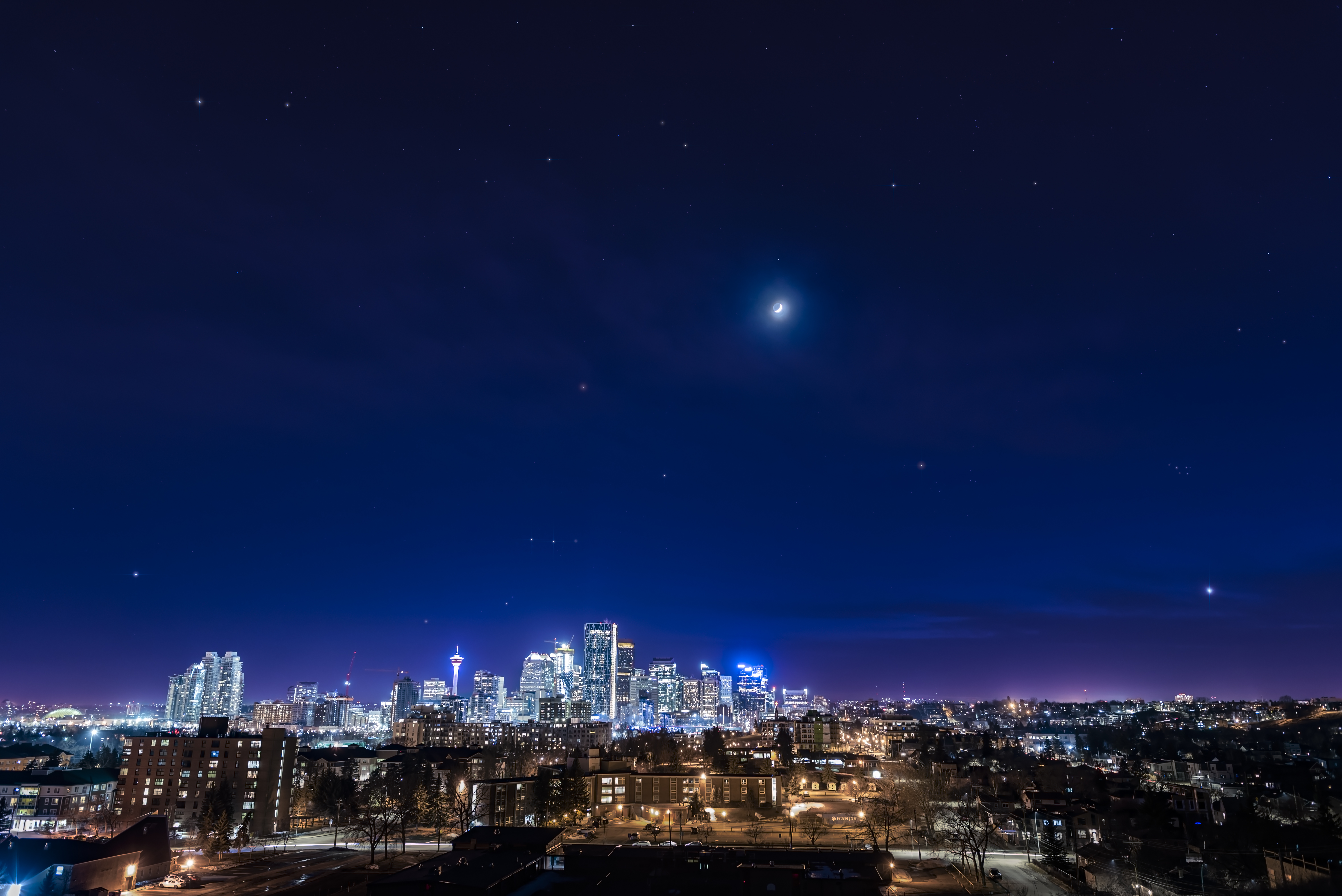 Venus (lower right), the Moon (center top), and a few bright stars — including those of the Pleiades and Orion — appear above the city of Calgary, Alberta, 
in this composite image meant to mimic how the human eye saw the scene. Even so, the photographer notes the camera picked up a few more stars than were visible to the naked eye (perhaps because of light pollution.)