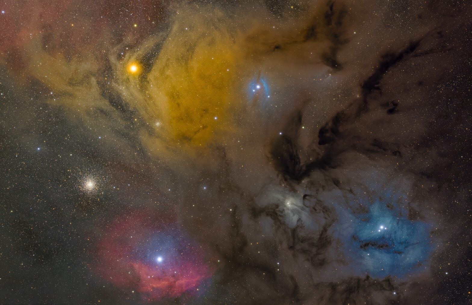 A photograph of the Rho Ophiuchi cloud complex. The image is dominated by swirling nebulae in various colors — yellows, blues, and hints of red and pink. At lower left lies the star Antares, enveloped by a golden yellow cloud. In the upper right is Rho Ophiuchi, surrounded by a blue reflection nebula. The background is filled with countless stars of varying brightness. Dark dust lanes create striking contrasts throughout the image, particularly in the center. The overall effect is a rich, colorful cosmic tapestry of gas, dust, and stars.
