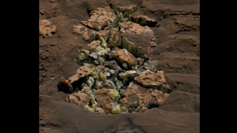An image of a pile of rocks on Mars. The soil around the rocks is a terra cotta color and the rocks in the middle of the image feature yellow crystals on the edges.