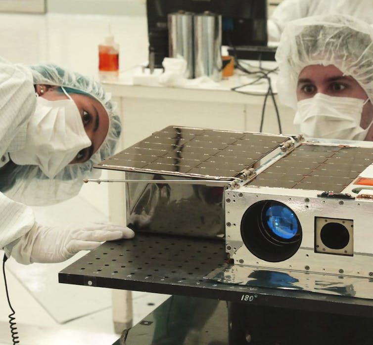 Two scientists wearing masks, gloves, head coverings and white clean suits work on an instrument in a laboratory.