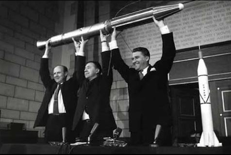 A black and white photo of three men holding a model of a cylindrical spacecraft over their heads.