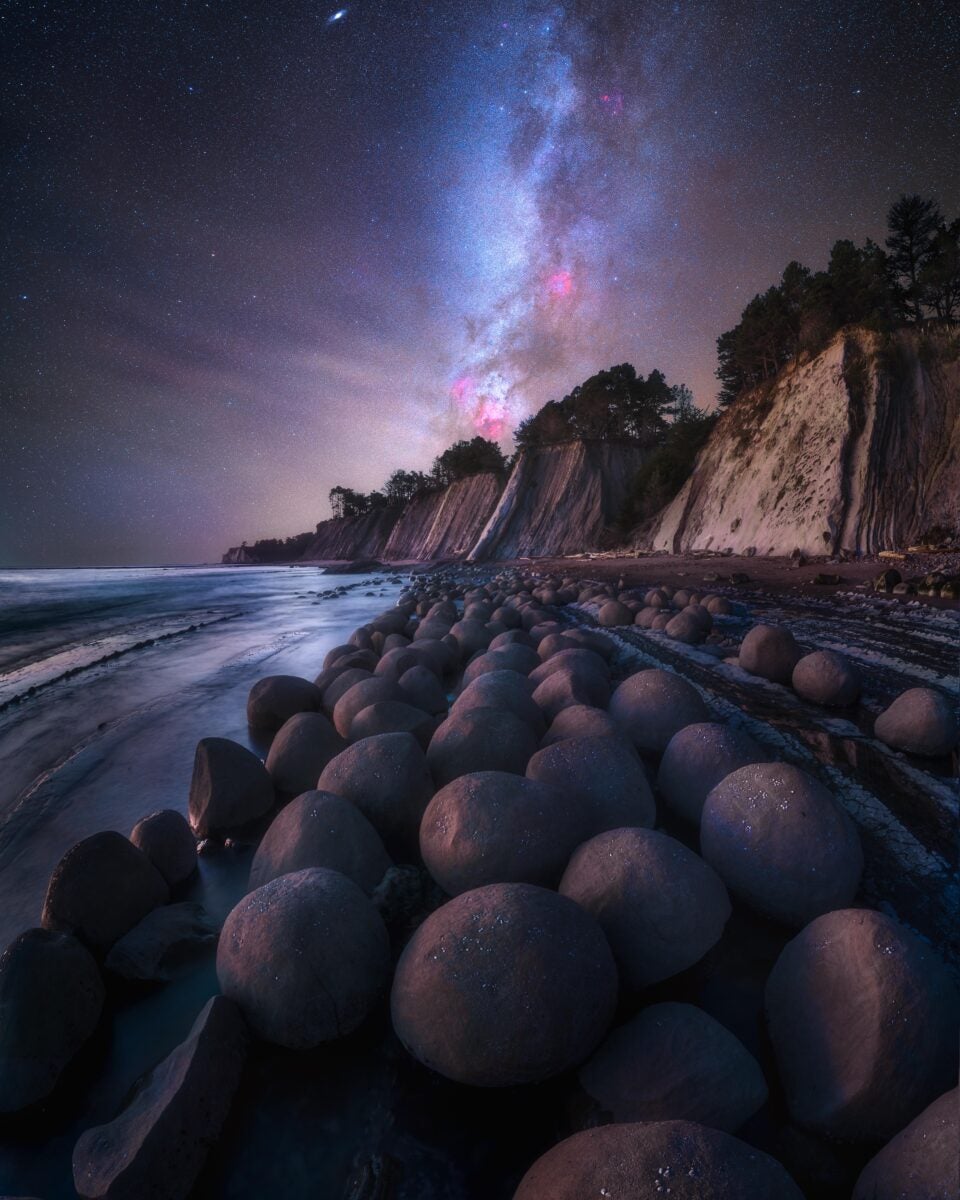 A nighttime photograph of a beach at low tide, revealing spherical rock formations that are typically underwater. In the foreground, smooth, rounded rocks are visible in low light, with waves gently lapping from the left. Coastal cliffs rise in the background, with trees silhouetted at the top. The sky dominates the upper portion of the image, with the Milky Way stretching at a slight angle from vertical across the frame. Colors in the sky include shades of pink, purple, and blue against the darker night sky. The composition creates a sense of depth, with the rocks in the foreground leading to the cliffs and then to the Milky Way, extending above and out of frame.